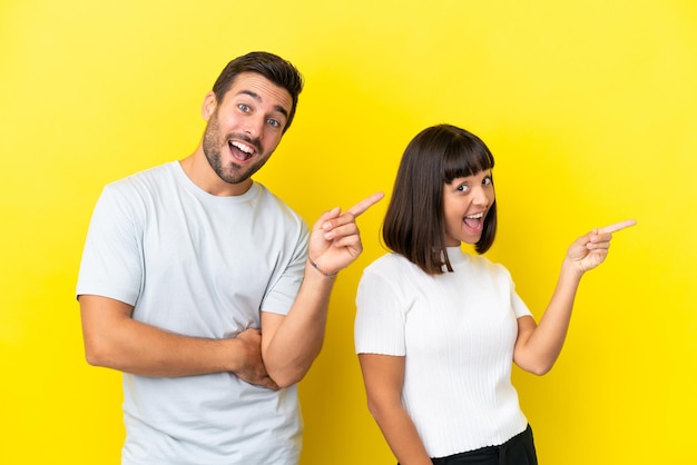 Young couple isolated on yellow background pointing finger to the side and presenting a product