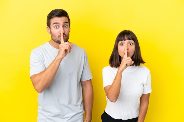 Young couple isolated on yellow background showing a sign of silence gesture putting finger in mouth