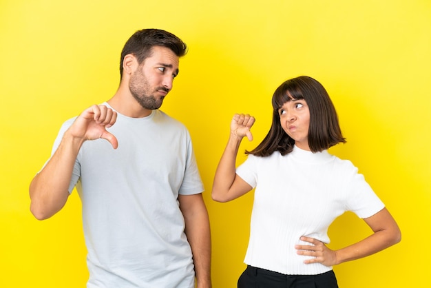 Young couple isolated on yellow background showing thumb down sign with negative expression