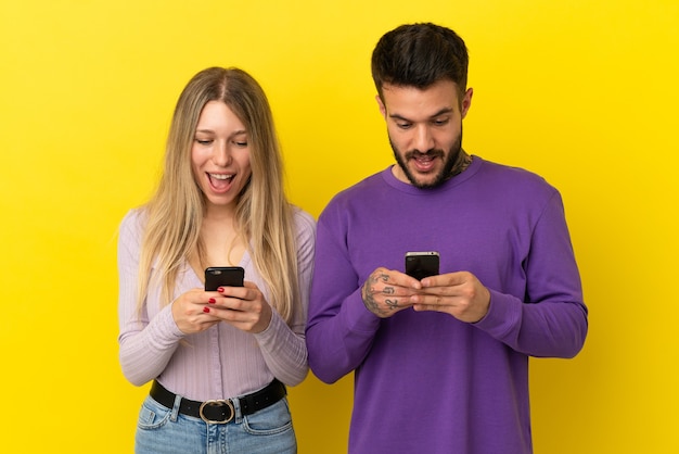 Giovane coppia su sfondo giallo isolato sorpreso e invia un messaggio o un'e-mail con il cellulare Foto Premium