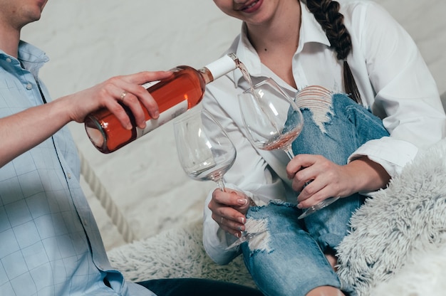 A young couple in jeans and shirts pours rose wine from bottle into glasses sitting on bed