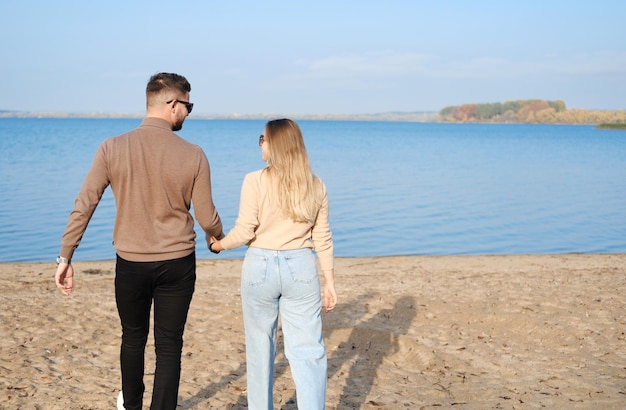 A young couple in jeans and sweaters hold hands and walk along the beach Back view Sunny weather