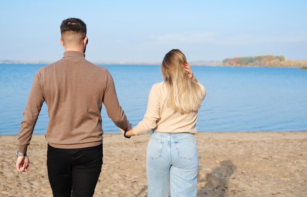 A young couple in jeans and sweaters hold hands and walk along the beach Back view