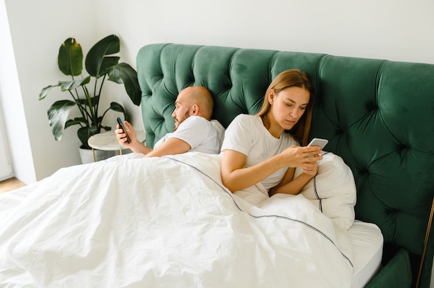 Photo young couple lie in bed with their phones and turned away from each other