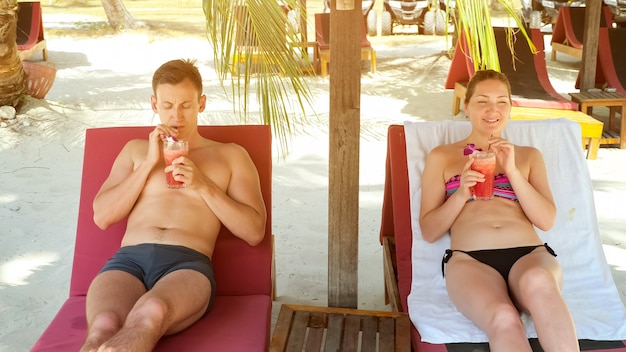 Young couple lies on sunbeds and drinks tasty beverages under shed enjoying holiday on tropical country beach closeup