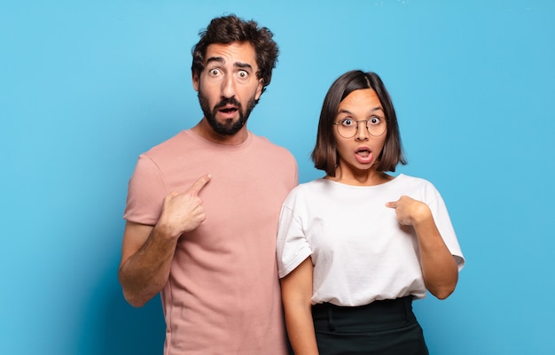 Young couple looking shocked and surprised with mouth wide open, pointing to self