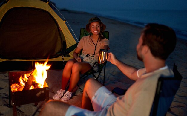 A young couple in love are sitting near the tent on folding\
chairs by the fire, drinking tea at night on the seashore.