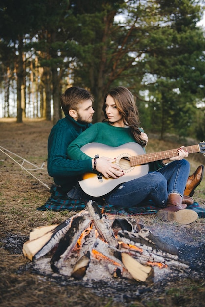 Young couple in love camping tourists sitting by a campfire\
against a tent in the woods playing guitar