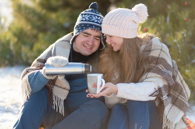 Young couple in love drink a hot drink from a thermos, sitting in the winter in the forest, tucked into warm, comfortable rugs, and enjoy nature. The guy pours a drink from a thermos in a cup