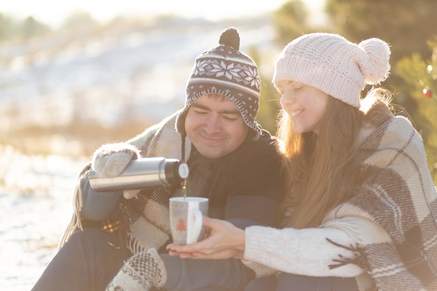 Photo young couple in love drink a hot drink from a thermos, sitting in the winter in the forest, tucked into warm, comfortable rugs, and enjoy nature