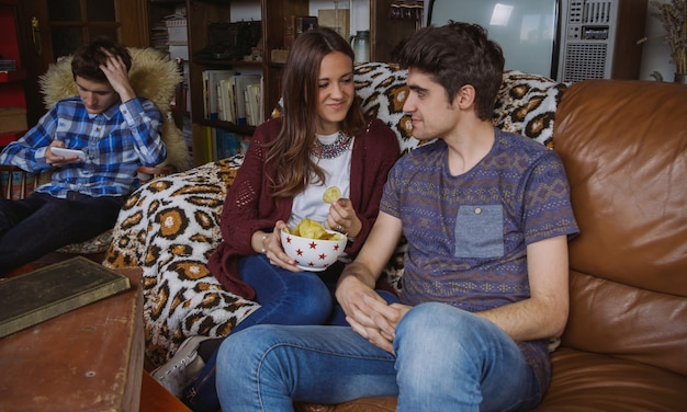 Young couple in love eating fried chips sitting on a sofa and their friend looking smartphone in a bored day at home. Teenager leisure time concept.