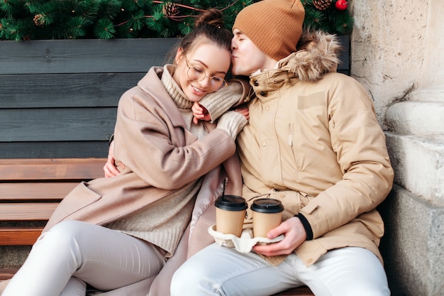 若いカップルの恋人は、冬に屋外で楽しく、一緒にコーヒーを飲みながらベンチに座っている