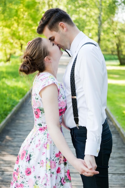 Photo young couple in love holding hands and looking each other in the eyes in summer park straight wooden path woman in dress and man wearing shirt with suspenders