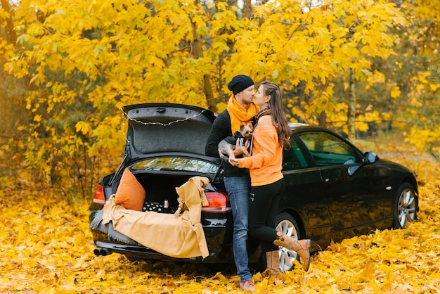 A young couple in love is sitting on the open trunk of a black car with their little dog in the autumn forest. Lovers kiss, and the dog looks at them