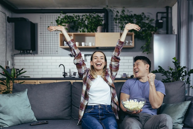 Молодая влюбленная пара мужчина и женщина вместе смотрят телевизор дома, радуясь и поедая попкорн счастливая многорасовая семья в новой квартире
