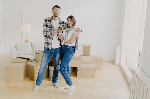 Photo young couple in love stand closely to each other embrace and holds favourite dog pose in empty room surrounded with cardboard boxes make family portrait in new apartment moving day concept