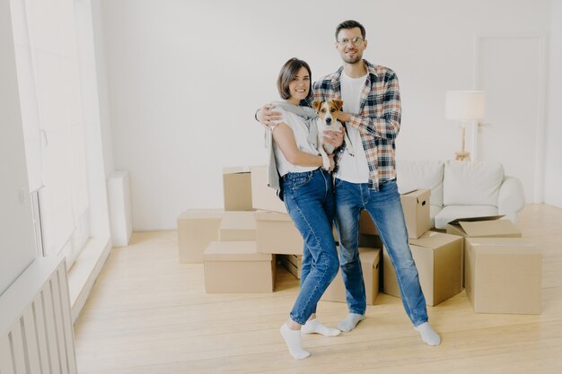 Young couple in love stand closely to each other, embrace and holds favourite dog, pose in empty room