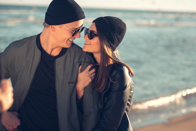 愛の若いカップルは海沿いを歩きます。春、秋。男はジャケットと帽子をかぶっています。帽子とスカーフと革のジャケットの女の子