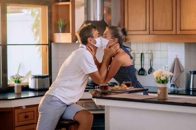 Giovane coppia innamorata indossa maschere protettive e si baciano in cucina, pandemie e sentimenti.