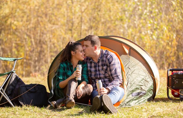 Young couple in love with thermos in hand kissing in front of the tent. Happy couple on vacation