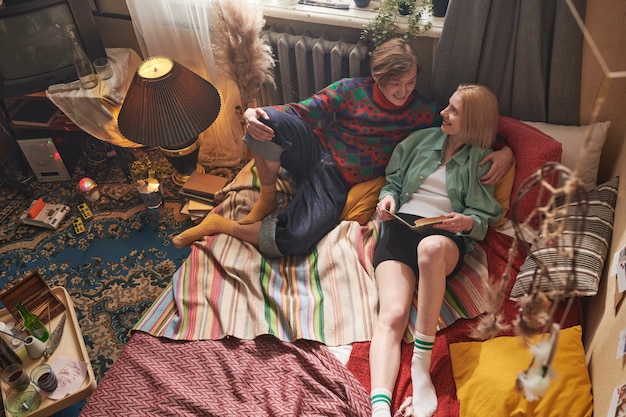 Young couple lying on bed embracing each other and reading a book in old-fashioned room at home