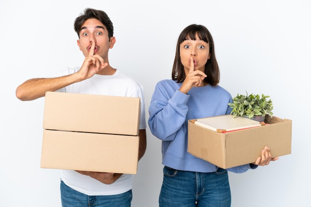 Young couple making a move while picking up a box full of things isolated on white background showing a sign of silence gesture putting finger in mouth