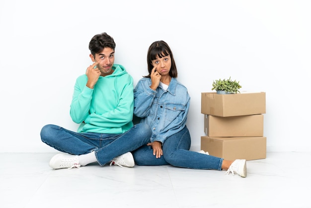 Молодая пара делает ход, поднимая коробку с вещами, сидящими на полу на белом фоне и смотрящими вперед