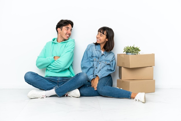 Молодая пара делает ход, поднимая коробку с вещами, сидящими на полу на белом фоне, с улыбкой оглядываясь через плечо
