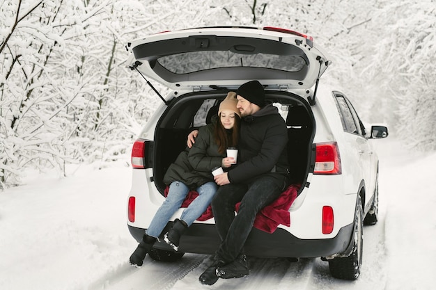 若いカップルの男と女が抱き合って冬の雪に覆われた森で車のトランクに座っています。