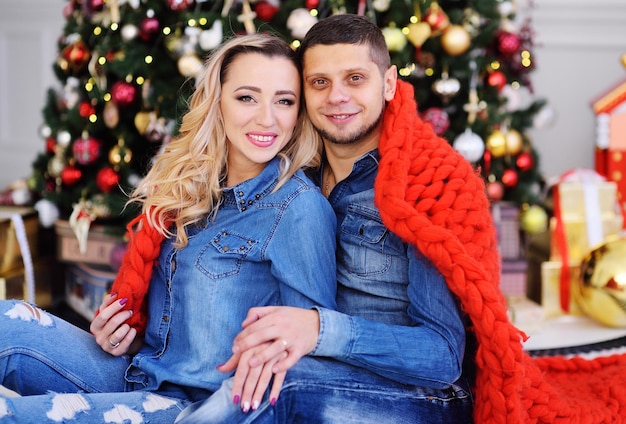 A young couple-a man and a woman in denim clothes covered with a red warm blanket and hugging smile on the background of Christmas trees and new year's decor.