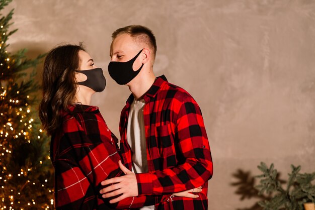 A young couple man and woman in disposable medical masks under a Christmas tree.