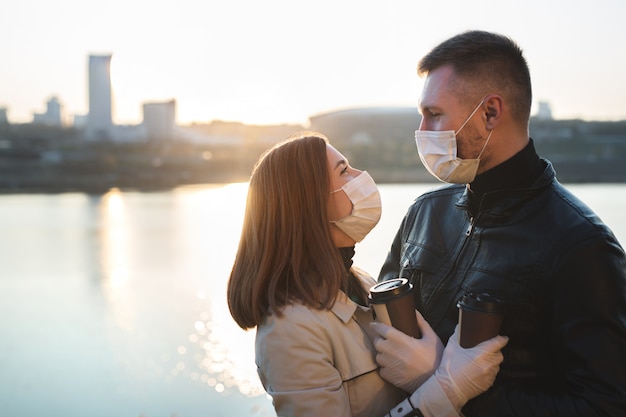 Photo young couple, a man and a woman in medical masks and gloves, drink coffee from disposable cups on the street and look at each other