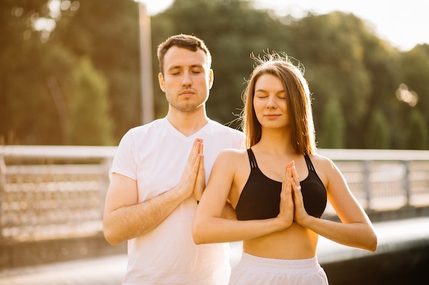 Young couple man and woman meditate together on sunset while standing, yoga on city lawn, summer evening, lifestyle