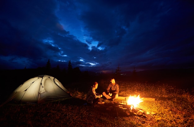 Young couple man and woman tourists enjoying at night camping in the mountains, sitting near burning campfire and illuminated tourist tent under beautiful evening cloudy sky