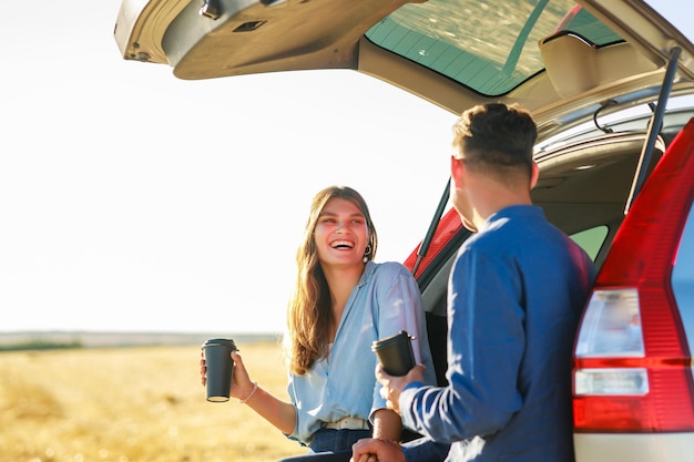 日没時に麦畑でコーヒーを飲むために停車する新しい車で一緒に旅行する若いカップルの男女