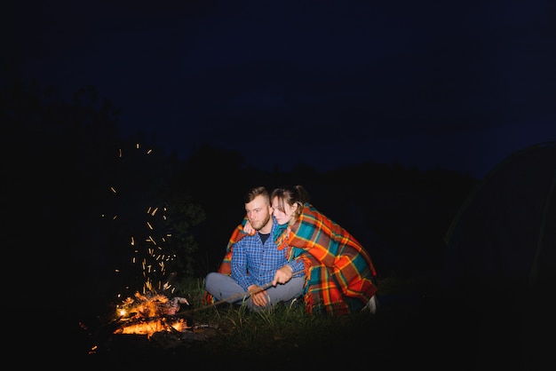 Young couple man and woman trevelers sitting near glowing tourist tent, burning campfire, on the top of mountain