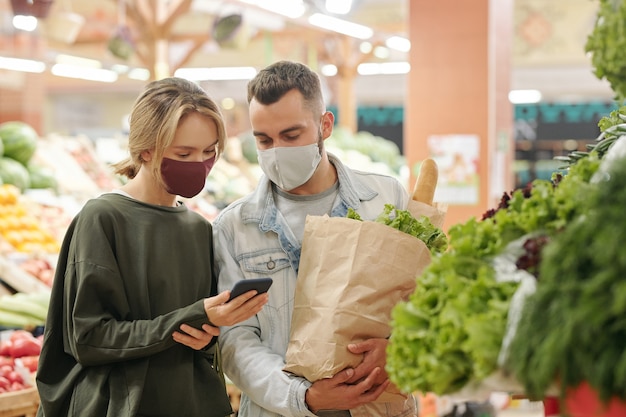 Young couple in masks standing at counter and checking fruit together before buying