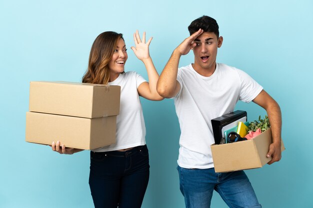 Young couple moving in new home among boxes on blue with surprise and shocked facial expression