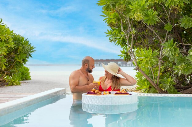 신혼부부는 신혼여행을 즐기고 수영장에서 떠다니는 접시와 함께 음료와 간식을 마르디브의 열대 섬 휴양지에서 즐기고 로맨틱한 데이트를위한 아침 식사를 즐기고 있습니다.