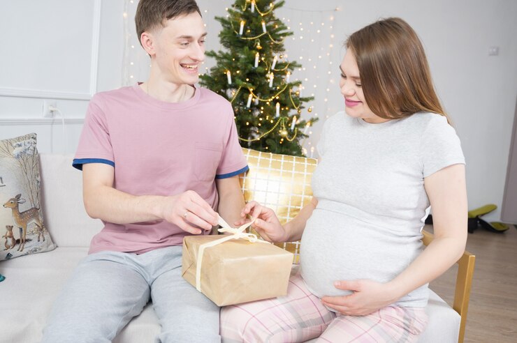 Young couple opening christmas present box, sitting on sofa with Christmas decoration in background.