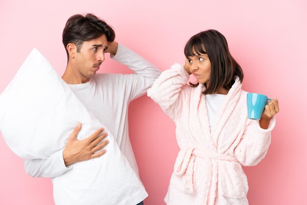 Young couple in pajamas isolated on pink background having doubts while scratching head