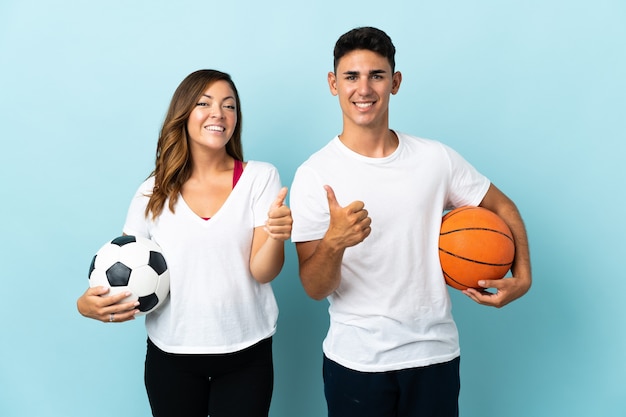 Young couple playing football and basketball on blue giving a thumbs up gesture with both hands and smiling