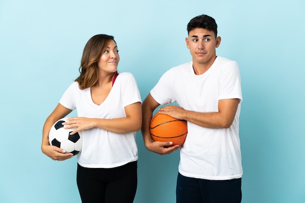 Молодая пара играет в футбол и баскетбол на синем, делая жест сомнения, поднимая плечи