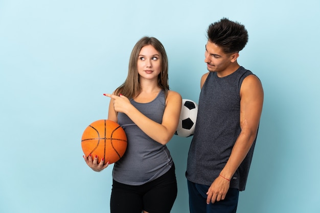 Молодая пара играет в футбол и баскетбол на синем, представляя идею, улыбаясь в сторону