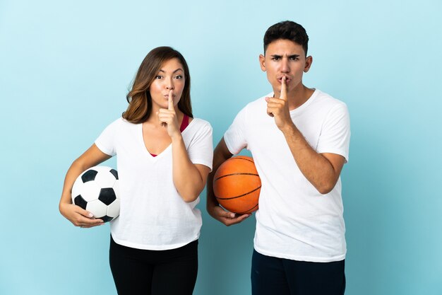 Young couple playing football and basketball on blue showing a sign of silence gesture putting finger in mouth