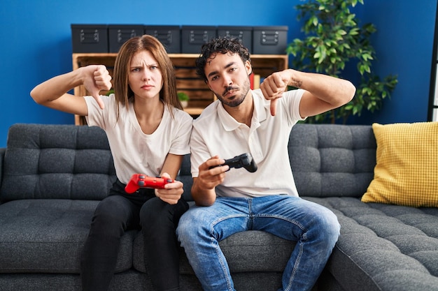 Young couple playing video game holding controller at home with angry face, negative sign showing dislike with thumbs down, rejection concept