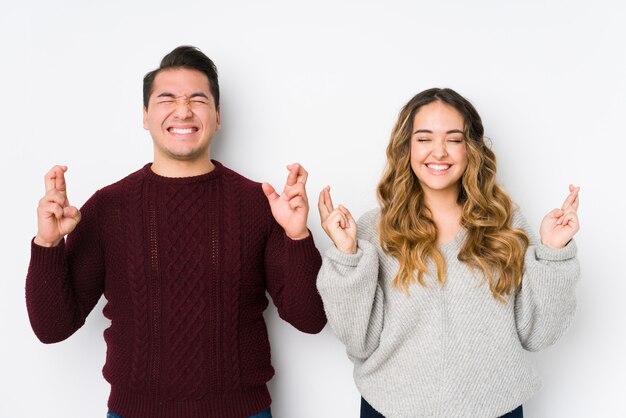 Young couple posing in a white wall crossing fingers for having luck