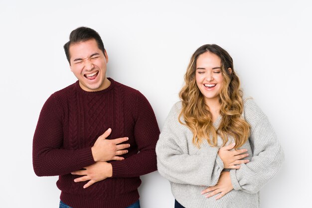 Young couple posing in a white wall laughs happily and has fun keeping hands on stomach.