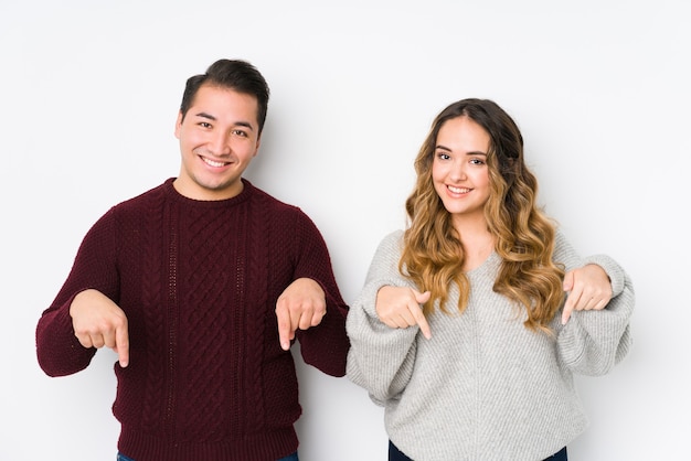 Young couple posing in a white wall points down with fingers, positive feeling.