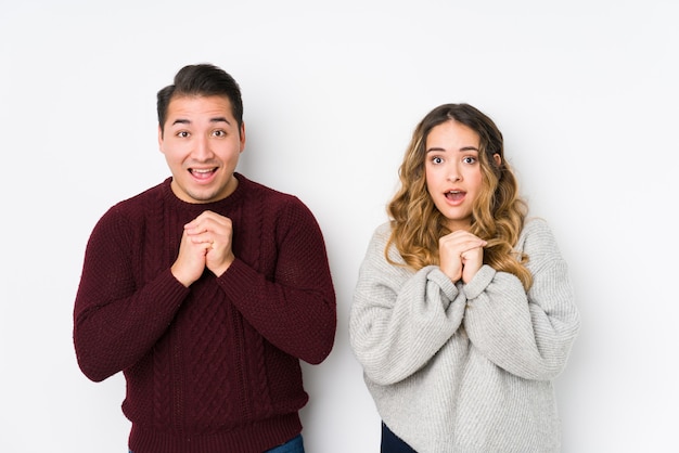 Young couple posing in a white wall praying for luck, amazed and opening mouth looking to front.